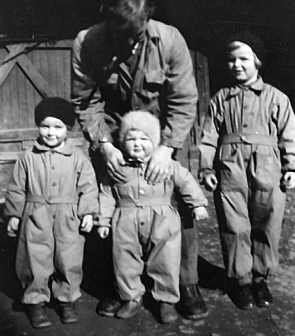 Black-and-white photograph: three children in snowsuits and knitted hats in front of a wooden building. An adult bends over the smallest child, holding it.