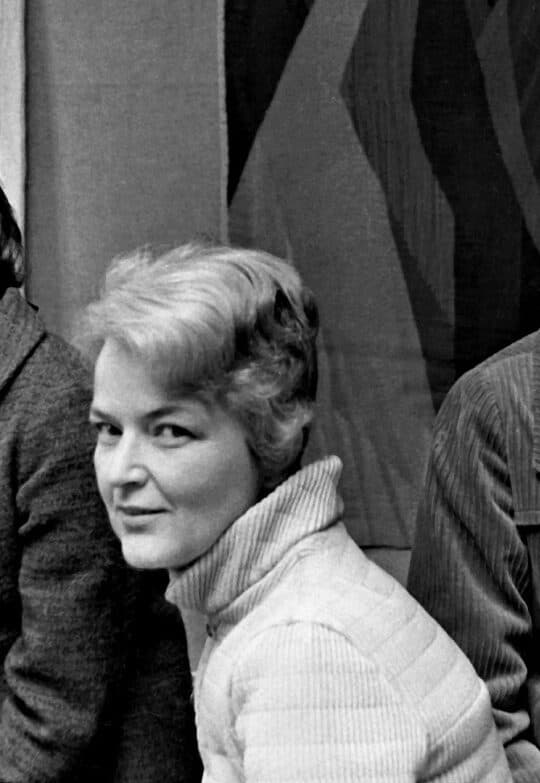 Svart-hvitt-fotografi av Karin Sundbye i lys, quiltet jakke og høyhalset genser. Bak henne henger et billedteppe med mørke, kantede former.