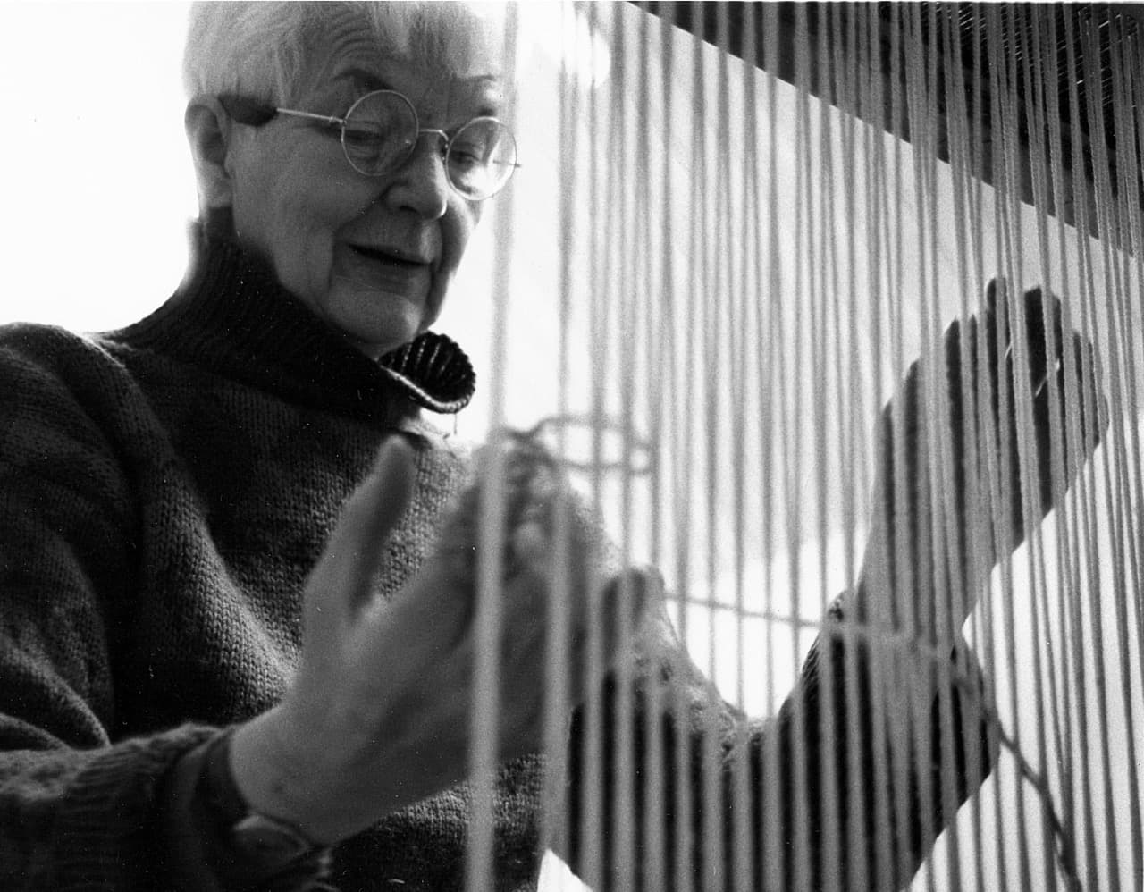 Svart-hvitt-fotografi: Karin ved vevstolen, sett gjennom de stramme renningstrådene, med blikket vendt ned mot hendene i arbeid.