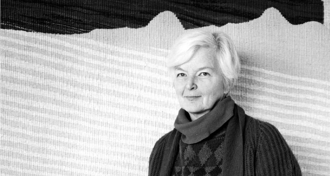 Svart-hvitt-fotografi: Karin foran et lyst, vevd teppe i brede vannrette striper med et mørkt, bølgende felt øverst.