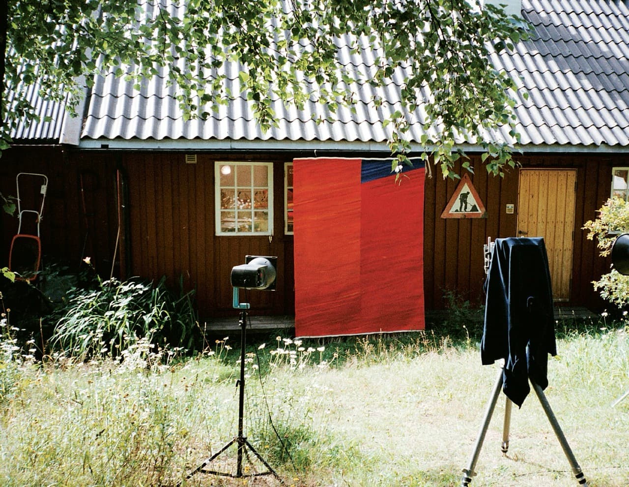 Fotografi av et rødt billedteppe hengt til fotografering på veggen av et rødmalt trehus, med lampestativ og kamerastativ i gresset foran.