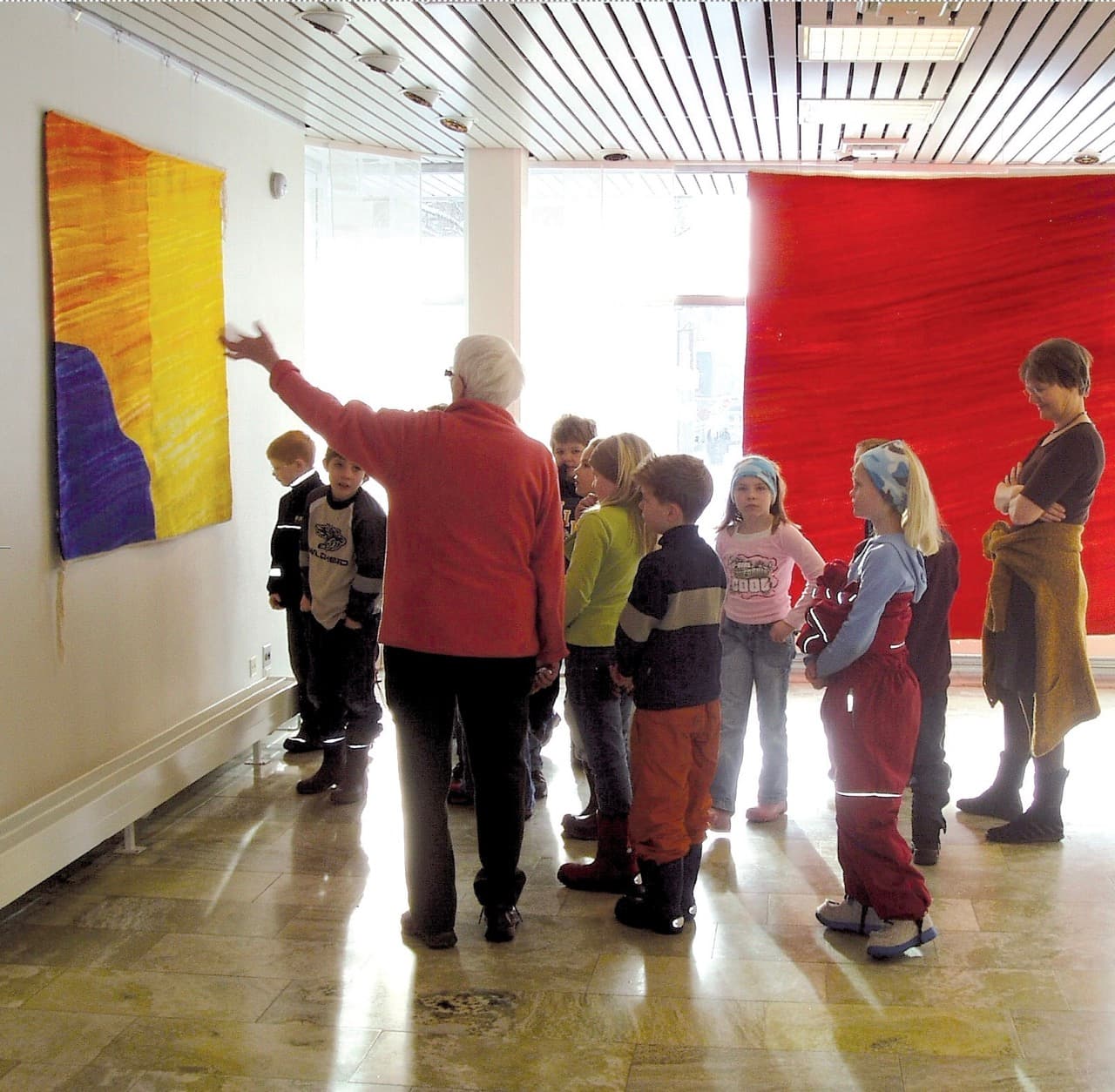 Fotografi fra en utstilling. En kvinne i rød genser peker på et gult og fiolett billedteppe for en gruppe skolebarn, med et stort rødt teppe i bakgrunnen.
