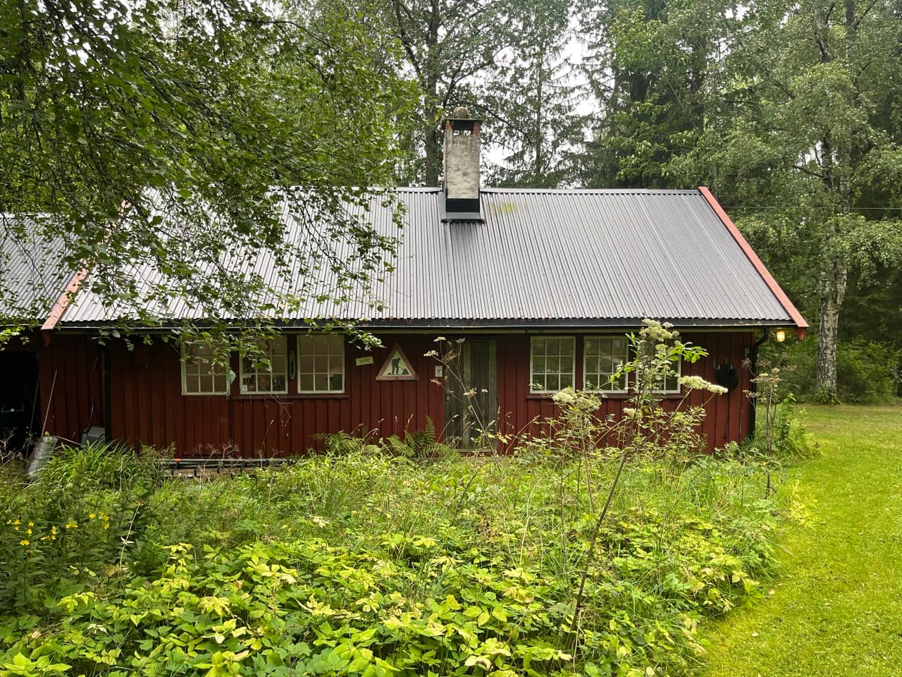 Fotografi av en lav, rødmalt bygning med bølgeblikktak og hvit skorstein, sett fra en gressplen. Høyt gress og bregner vokser tett foran fasaden, med granskog bak.