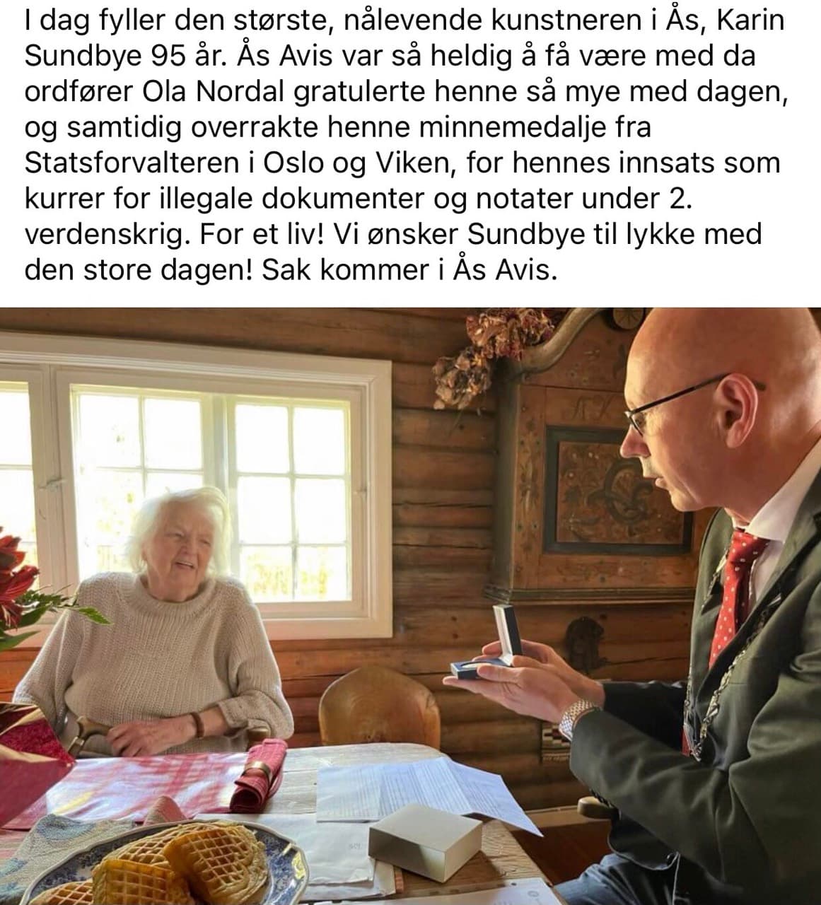 Skjermbilde av et innlegg fra Ås Avis om at Karin Sundbye fyller 95 år og mottar minnemedalje for sin innsats som kurer under andre verdenskrig. Under teksten et foto der ordføreren rekker fram medaljeesken over et kjøkkenbord.