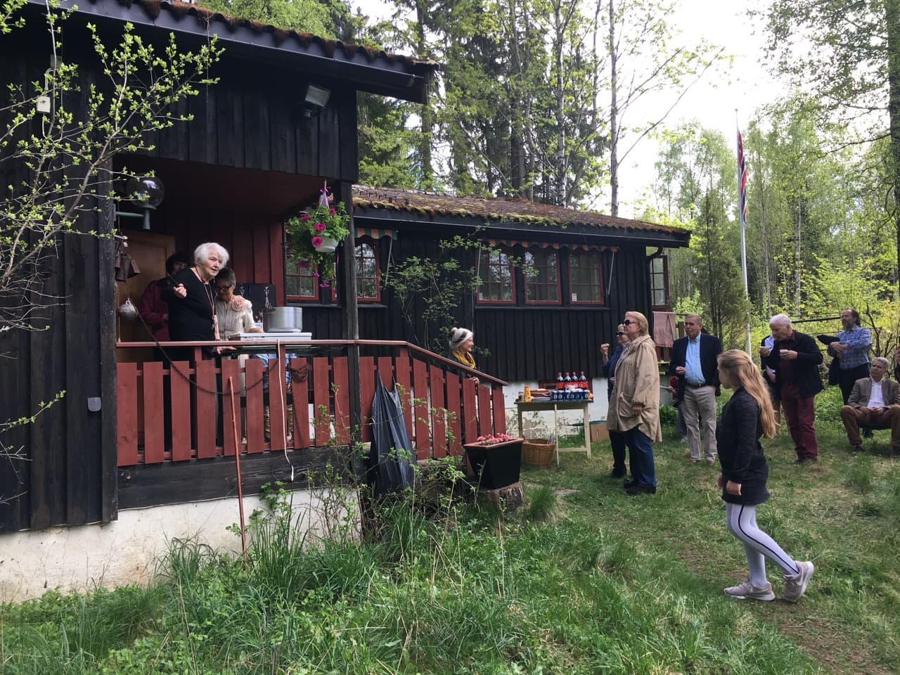 Photograph from a gathering outside a dark-stained wooden house. An elderly woman stands speaking from a veranda with a red railing, while guests stand on the grass in front, with spruce forest and a flagpole behind.