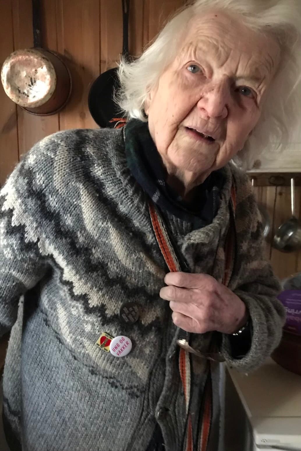 Portrettfoto av Karin Sundbye i strikkejakke på kjøkkenet, med kobberkjeler hengende på veggen bak.